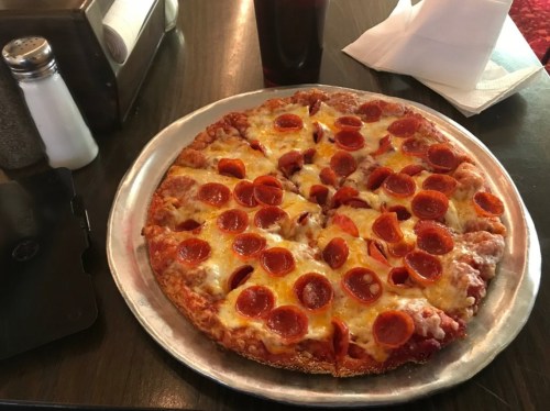 A pepperoni pizza on a metal tray, with melted cheese and a side of condiments, next to a glass of soda.