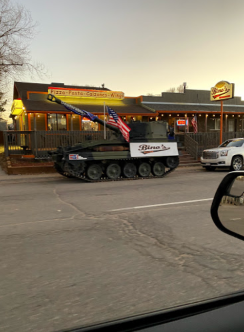 A tank with flags parked outside a restaurant named "Bino's," which serves pizza, pasta, calzones, and wings.