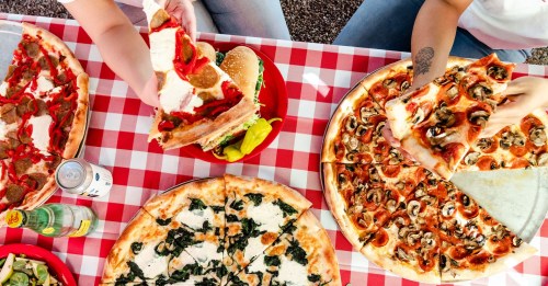 A table with various pizzas, a burger, and drinks, set on a red and white checkered tablecloth.