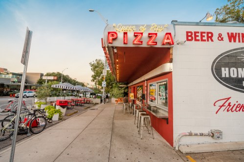 A pizza restaurant with a bright sign, outdoor seating, and nearby bicycles on a sunny day.