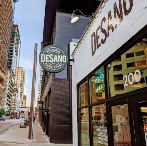 Sign for Desano Pizzeria Napoletana on a city street, with buildings and a clear sky in the background.