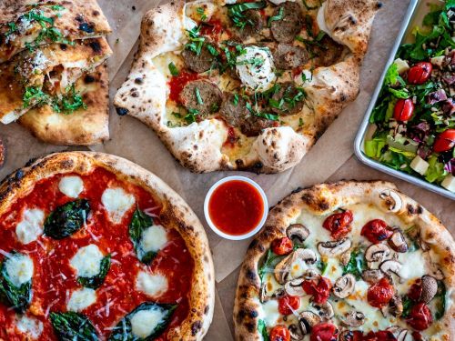 A variety of pizzas on a table, including margherita, meat, and veggie, with a side of marinara sauce and a salad.