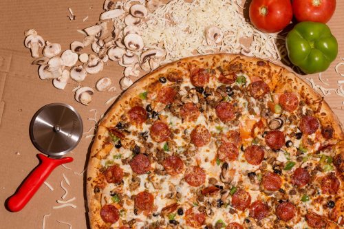 A large pepperoni pizza with toppings, surrounded by sliced mushrooms, tomatoes, green peppers, and cheese shreds.
