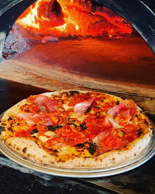 A freshly baked pizza topped with tomato sauce, cheese, and slices of prosciutto, with a wood-fired oven in the background.