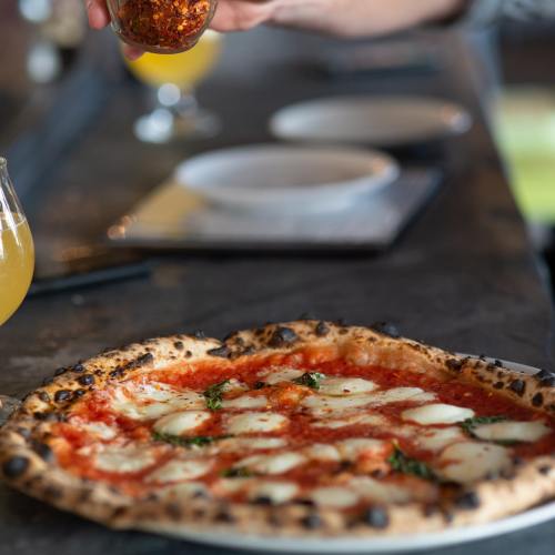 A close-up of a pizza with mozzarella and basil, with someone sprinkling red pepper flakes in the background.