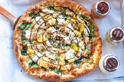 A freshly baked pizza topped with chicken, artichokes, spinach, and herbs, served with seasoning jars on the side.