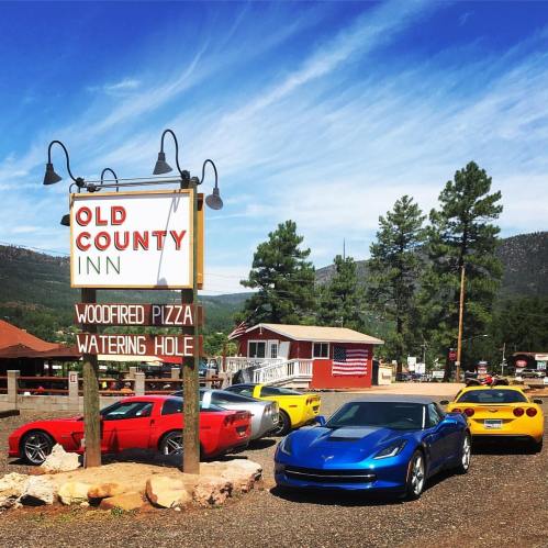 A row of colorful sports cars parked outside the Old County Inn, surrounded by trees and a clear blue sky.