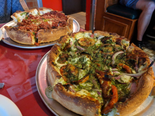Two deep-dish pizzas on metal trays: one with meat and cheese, the other topped with vegetables and pesto.
