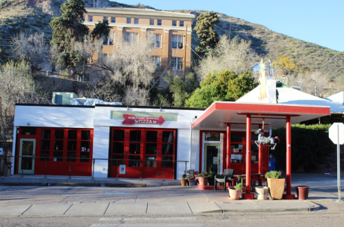A colorful pizza shop with large windows, surrounded by trees and a hillside, featuring a vintage gas station design.