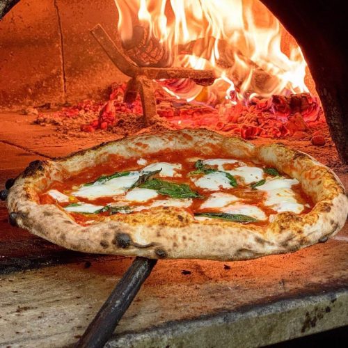 A freshly baked pizza with mozzarella and basil, emerging from a wood-fired oven with flames in the background.