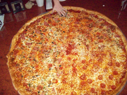 A large, round pizza topped with pepperoni, vegetables, and cheese, with a hand reaching for a slice.