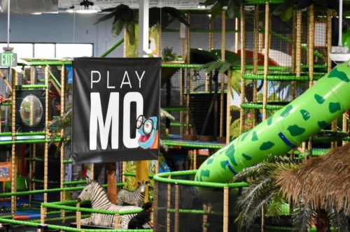 A colorful indoor play area featuring a large slide, jungle-themed decor, and a sign that says "PLAY MO."