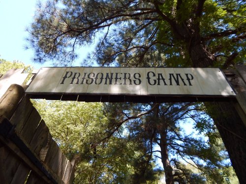 Sign reading "PRISONERS CAMP" above a wooden entrance, surrounded by tall trees and natural greenery.
