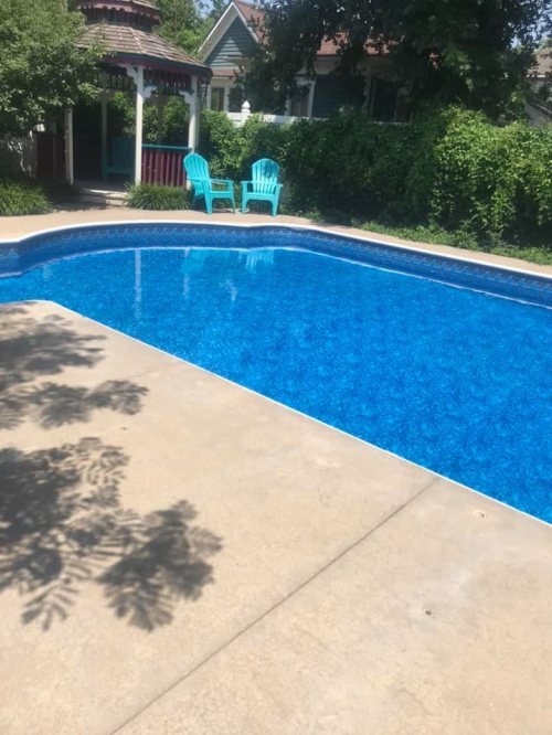 A clear blue swimming pool surrounded by a concrete deck, with two turquoise chairs and a gazebo in the background.