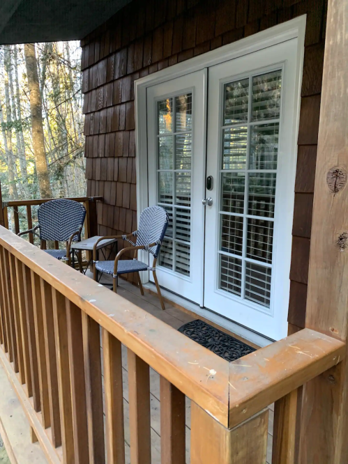 A wooden porch with two chairs and a welcome mat in front of double glass doors, surrounded by trees.
