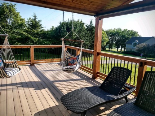 A wooden deck with hanging chairs and a lounge chair, surrounded by trees and a clear sky.