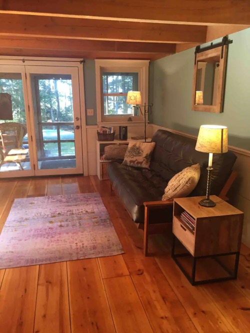 Cozy living room with a futon, wooden floor, a rug, and a door leading to a patio surrounded by trees.