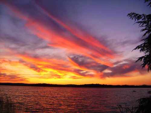 Vibrant sunset over a calm lake, with colorful clouds reflecting in the water and silhouettes of distant hills.