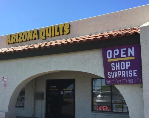 Storefront of Arizona Quilts with a purple "OPEN" sign and "SHOP SURPRISE" banner under a clear blue sky.