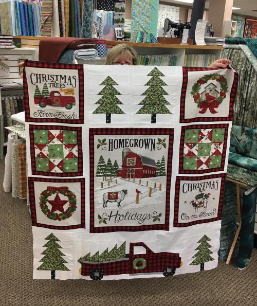 A person holds a festive quilt featuring Christmas trees, wreaths, and a farm scene with the text "Homegrown Holidays."