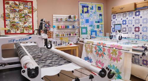 A quilting studio featuring a longarm quilting machine, colorful quilts on the walls, and organized sewing supplies.
