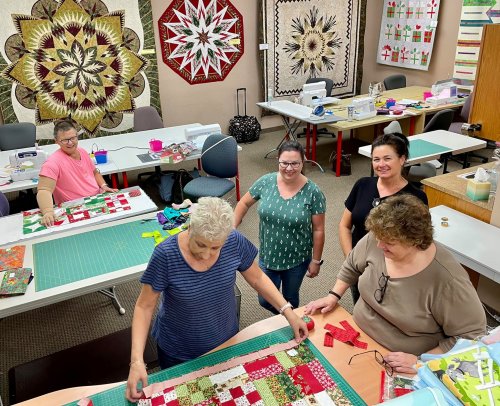 Five women work on quilting projects in a bright, organized sewing room with colorful quilts displayed on the walls.