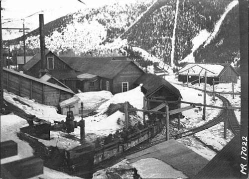 Historic black-and-white photo of a snowy mining town with buildings and a train track in a mountainous landscape.