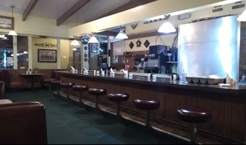 A cozy diner interior featuring a long counter with stools, booths, and warm lighting.