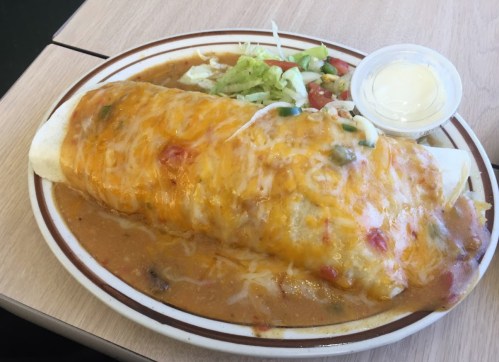 A large cheese-covered burrito on a plate with sauce, accompanied by lettuce and a small cup of dressing.