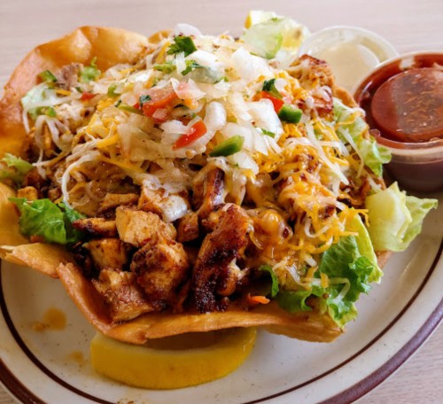 A colorful taco salad in a crispy shell, topped with grilled chicken, cheese, lettuce, and salsa on the side.