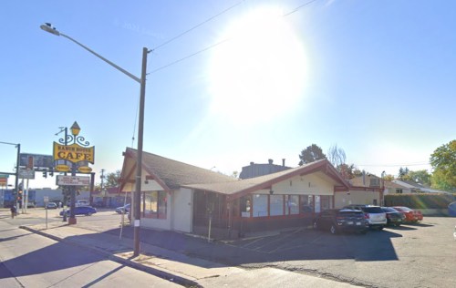 A sunny view of a cafe building with a sign, parked cars, and a clear blue sky in the background.