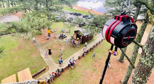 Aerial view of a group of people gathered outdoors near a lake, with a zip line pulley in the foreground.