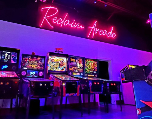 A vibrant arcade with neon "Reclaim Arcade" sign and several pinball machines lined up against a colorful wall.