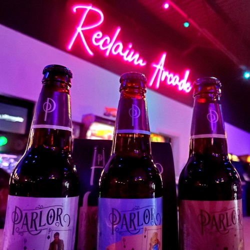 Three bottles of Parlor beer in front of a neon sign that reads "Reclaim Arcade," with colorful lights in the background.