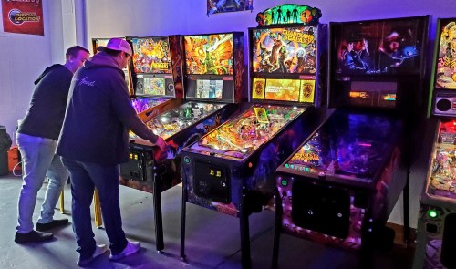 Two people playing pinball machines in a brightly lit arcade, surrounded by colorful game graphics.