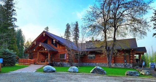 A rustic log cabin surrounded by trees, featuring a stone foundation and a grassy area with large boulders.