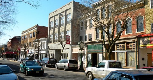 A street view featuring historic buildings, shops, and parked cars under a clear blue sky.