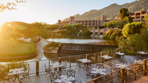 A scenic view of a resort with outdoor dining, a lake, and mountains at sunset.