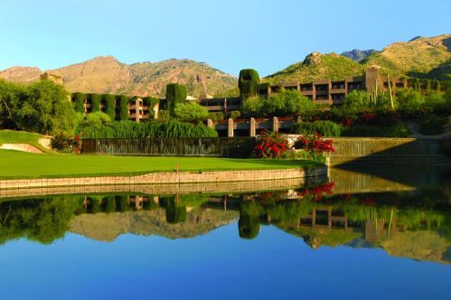 A serene golf course with lush greenery and mountains, reflecting in a calm lake under a clear blue sky.