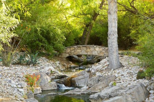 A serene landscape featuring a stone bridge over a gentle stream, surrounded by lush greenery and rocky terrain.
