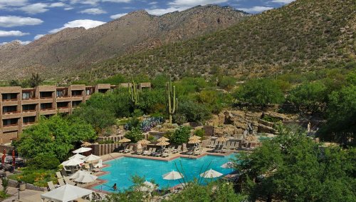 A scenic view of a resort pool surrounded by lush greenery and mountains under a partly cloudy sky.