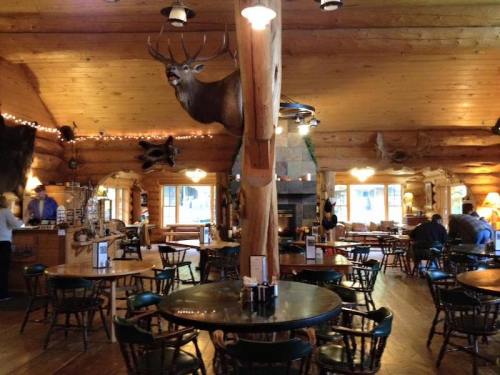 Cozy log cabin interior with wooden tables, a fireplace, and mounted deer heads on the walls. Patrons dining and socializing.