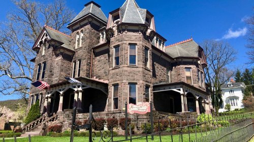 A large, historic stone mansion with multiple gables, surrounded by greenery and blue skies. Flags adorn the entrance.