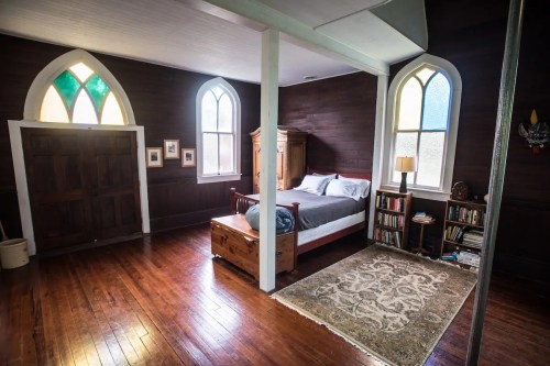 Cozy bedroom with wooden floors, arched windows, a bed, bookshelf, and decorative rug. Warm, inviting atmosphere.