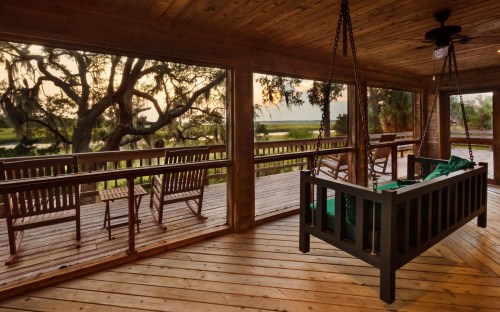 A cozy porch with a swing, rocking chairs, and a view of trees and a serene landscape at sunset.