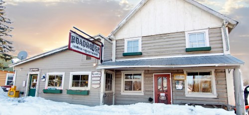 A rustic wooden building with a sign reading "Roadhouse," surrounded by snow and a colorful sunset sky.