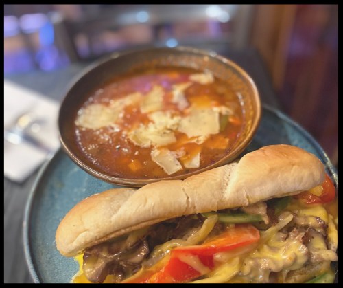 A plate featuring a sandwich with melted cheese and vegetables alongside a bowl of hearty soup.