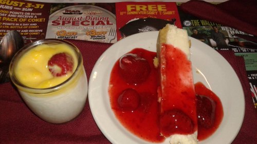 A dessert plate featuring a slice of cheesecake topped with strawberry sauce and fresh strawberries, alongside a cup of mango pudding.