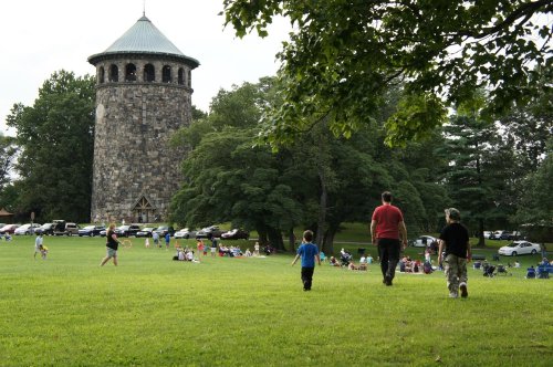 A grassy park with a stone tower in the background, where people gather for an outdoor event. Two children walk ahead.