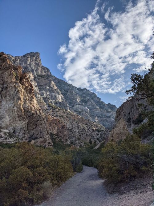 A winding path through rocky terrain, flanked by steep mountains and a blue sky with scattered clouds.